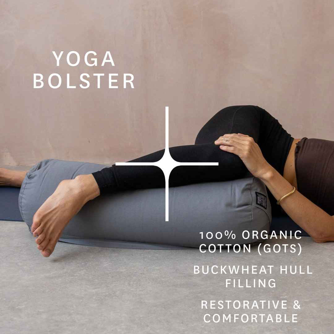 A person lying on a gray yoga bolster pillow in a studio setting, with text reading 'YOGA BOLSTER' and product info - organic cotton and buckwheat hull filling.