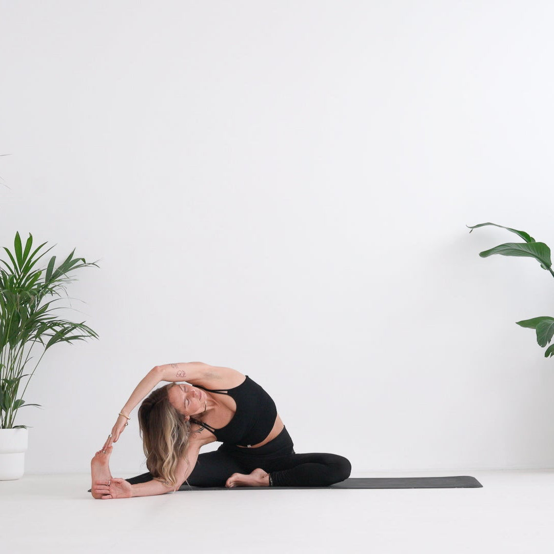 A woman in a black activewear set performs a side stretch yoga pose on a black mat in a bright, minimalist room with potted green plants on both sides.