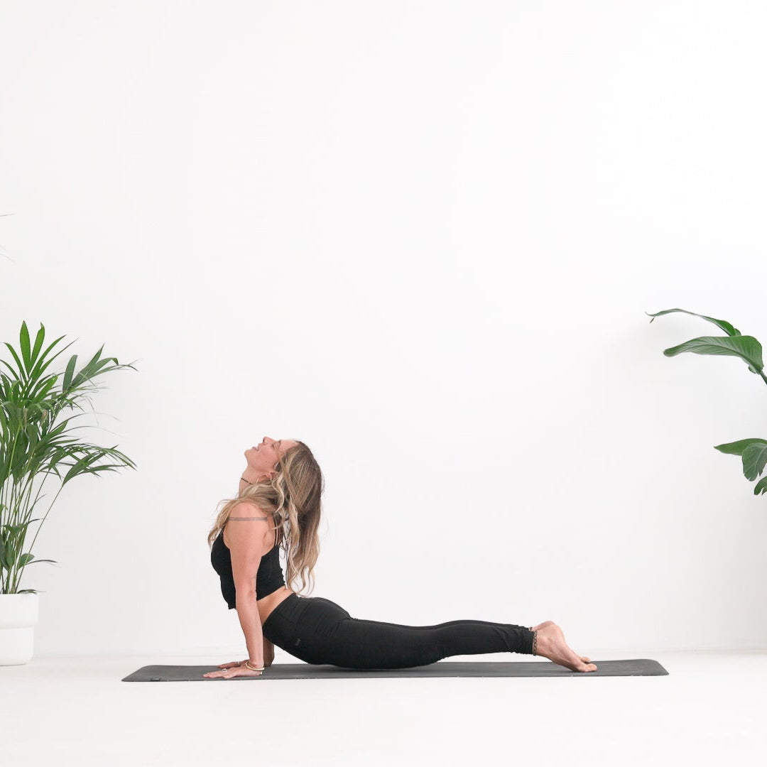 A woman does a yoga pose on a premium Movement mat in a bright, minimalist room with potted plants on both sides.